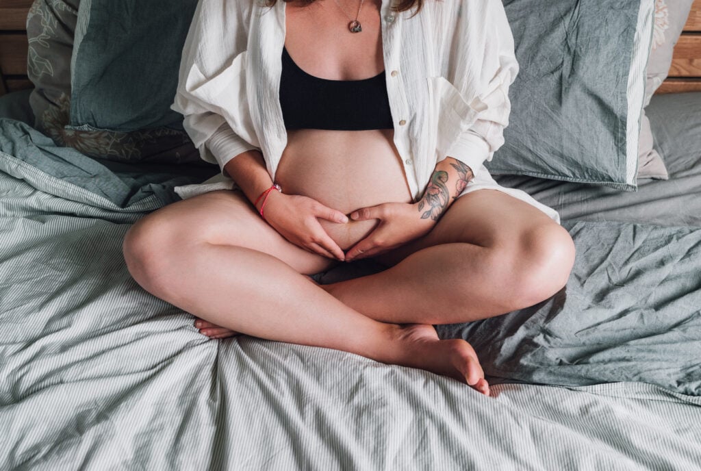 Young pregnant woman with tattooed hand sitting cross-legged tender touching belly on the bed in the bedroom in early morning time. Women's health, happy pregnancy, and calm mental mood concept image.
