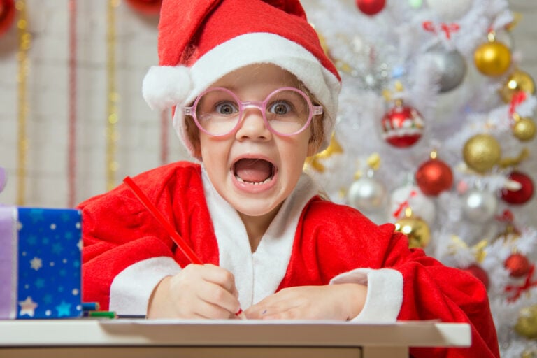 Child screams while writing and wearing a Santa hat by a Christmas tree