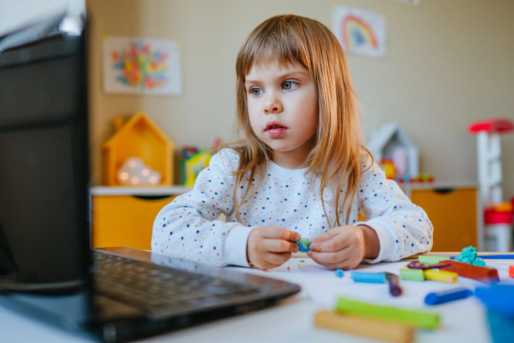 Little girl molding colorful clay watching online learning lesson on the laptop indoor. Distance home learning concept.