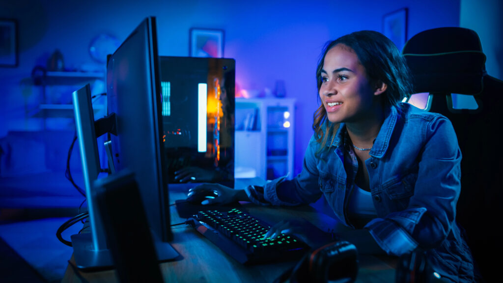Beautiful and Excited Black Gamer Girl is Playing First-Person Shooter Online Video Game on Her Computer. Room and PC have Blue Neon Led Lights. Cozy Evening at Home.