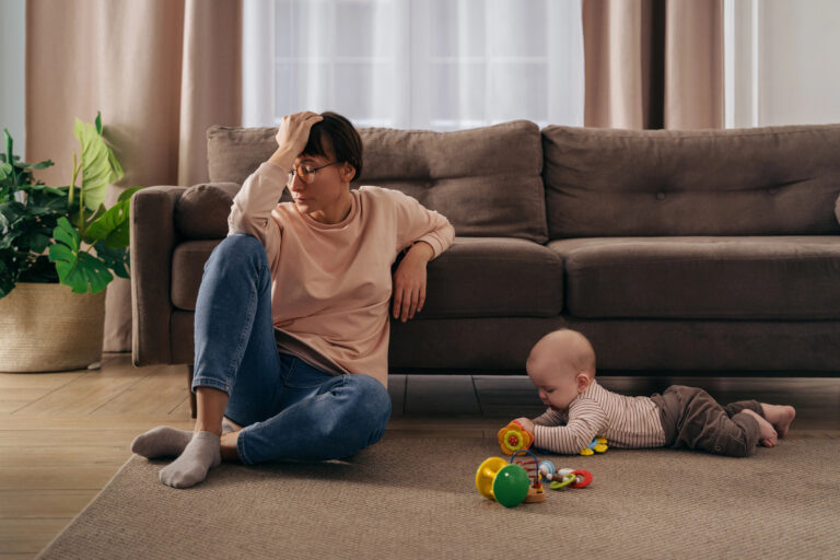 Young tired woman suffering from postnatal depression, holding her head with hand, sitting on floor in living room near her toddler baby playing with toys. New mom tired from motherhood difficulties