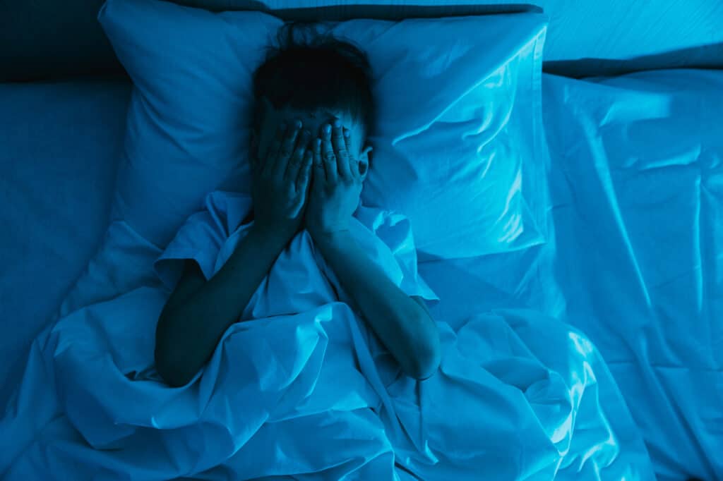 Small child boy lies in bed in dark night and covers his face with his hands in fear, afraid of nightmares and terrible dreams in children.