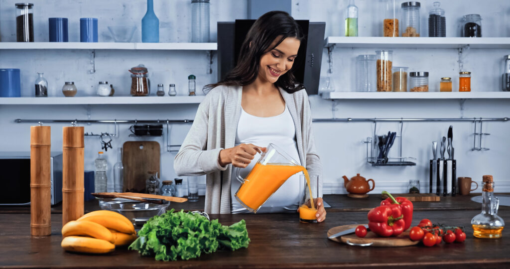 Smiling pregnant woman pouring orange juice near organic vegetables in kitchen