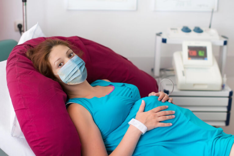 Pregnant patient in face mask in a hospital at doctor visit during coronavirus outbreak. Mother giving birth to baby in covid-19 lockdown. Pregnancy examination. Expecting woman in labor and delivery.